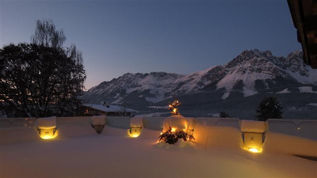 A snow-covered landscape with mountains in the background at dusk. Soft lights shine in the snow, creating a cozy atmosphere.