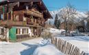 A traditional wooden house in the snow with a clear blue sky in the background. The surroundings are idyllic and peaceful, surrounded by mountains and a wooden fence.
