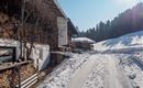 A snowy path leads alongside traditional wooden houses. The surroundings are quiet and surrounded by snow-covered mountains.