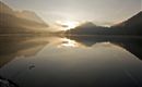 Ein ruhiger See mit spiegelndem Wasser und sanften Bergformen im Hintergrund. Die Morgensonne strahlt durch den Nebel und schafft eine friedliche Atmosphäre.