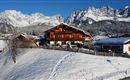A charming wooden house sits on a snow-covered hill. In the background, majestic mountains stretch under a clear blue sky.
