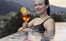 Eine Frau entspannt im Infinity-Pool und hält ein Cocktailglas mit einer Zitronenscheibe. Im Hintergrund sind sanfte Hügel und eine dramatische Wolkenkulisse zu sehen.