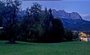 A calm landscape with a large tree and a house in the foreground. In the background, impressive mountains rise under an evening sky.