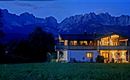 A modern house at dusk, surrounded by majestic mountains. The soft lights of the house shimmer in the serene landscape.