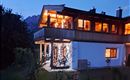 A modern house with cozy lights that shines in the twilight. In the background, gentle hills and a clear evening sky can be seen.