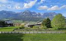 A picturesque landscape with green meadows and an impressive mountain range in the background. The sky is clear and blue with some clouds.