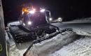 Ein Pistenraupe arbeitet nachts auf einem schneebedeckten Weg. Die Lichter des Fahrzeugs leuchten in der Dunkelheit.