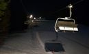 Eine Skiliftanlage bei Nacht auf einer verschneiten Piste. Die Umgebung ist düster, mit wenigen Lichtquellen in der Ferne.