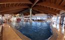 Ein großes Hallenbad mit klarem Wasser und Holzdecke. Umgeben von gemütlichen Liegen und Sitzbereichen für Gäste.