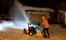A person is operating a snow blower in a snowy environment at night. The snow is being blown into the air, and a brightly lit house can be seen in the background.
