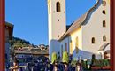 Eine Prozession in traditioneller Tracht geht an einer Kirche vorbei. Die Sonne scheint und der Himmel ist blau.