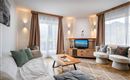 A modern living room with a cozy sofa, a wooden table, and a television. Large windows let in a lot of light and give the room an inviting atmosphere.