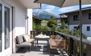 A cozy terrace with a table and chairs, surrounded by a beautiful mountain landscape. The sun is shining and the sky is blue.