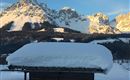 A cozy wooden house with a snow-covered roof. In the background, majestic mountains rise under a clear sky.