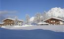A beautiful winter landscape with snow-covered huts and tall mountains in the background. The clear blue sky completes the idyllic scene.