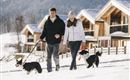 Ein Paar spaziert mit zwei Hunden durch einen verschneiten Bereich. Im Hintergrund sind moderne Holzhäuser und Berge zu sehen.