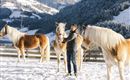Ein Mann steht im Schnee und streichelt ein Pferd. Im Hintergrund sind weitere Pferde und eine verschneite Berglandschaft zu sehen.