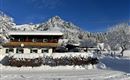 A picturesque wooden house in the snow, surrounded by snow-covered trees and mountains. The clear blue sky complements the winter landscape.