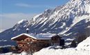 A charming wooden house in the snow with impressive mountains in the background. The landscape is wintery and peaceful.