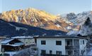 A picturesque winter landscape with snow-covered mountains in the background. In the foreground stands a modern residential house.