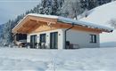 Ein modernes Haus im Schnee, umgeben von schneebedeckten Bäumen. Die Berge im Hintergrund bieten eine malerische Kulisse.