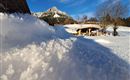 A snowy landscape with a view of a mountain in the background. In the foreground lies a slope with fresh snow, next to it is a cozy cabin.