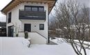 A modern house with a large balcony, surrounded by snow. In the background, bare trees are visible.