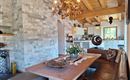 Een gezellige eetruimte met een grote houten tafel en zitplaatsen. De muren zijn bekleed met natuursteen, en er is een lichte, uitnodigende keuken op de achtergrond.
