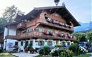 A traditional alpine house with wooden cladding and many flower balconies. The surroundings are green and sunny, with mountains in the background.