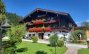 A traditional alpine house with blooming plants and a well-maintained garden. In the background, green mountains and a blue sky are visible.