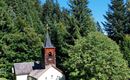 A small church surrounded by tall trees and green meadows. The sky is clear and blue.