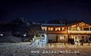 Ein gemütliches Chalet im Schnee bei Nacht. Im Hintergrund sind Berggipfel zu sehen, während die Lichter des Hauses strahlen.