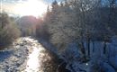 Ein klarer Fluss fließt durch eine verschneite Landschaft. Sonnenstrahlen scheinen auf das Wasser und die Bäume sind mit Schnee bedeckt.