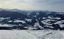 Eine winterliche Berglandschaft mit schneebedeckten Hügeln und mächtigen Bergen im Hintergrund. Der Himmel ist klar und die Sonne scheint.