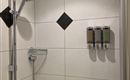 Een moderne douche met witte tegels en een douchekop. Aan de muur zijn drie dispensers voor shampoo en douchegel aangebracht.