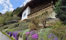 Ein hübsches Haus mit einem Balkon, umgeben von bunten Blumen. Im Hintergrund sind grüne Hügel und ein blauer Himmel zu sehen.