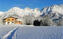 A cozy house is situated in a snow-covered landscape. In the background, majestic mountains rise under a blue sky.