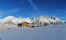 A beautiful wooden house is situated in a snow-covered landscape. In the background, impressive mountain peaks and a clear blue sky can be seen.