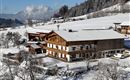 Ein malerisches Chalet im Schnee mit Bäumen und Bergen im Hintergrund. Die Landschaft ist ruhig und winterlich.
