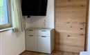 Ein schlichtes Zimmer mit einem Fernseher an der Wand und einem Holztür. Der Boden ist aus Holz und es gibt einen kleinen Schrank unter dem Fenster.