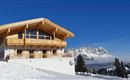 A modern chalet in the mountains, surrounded by snow. In the background, majestic mountains and a blue sky can be seen.