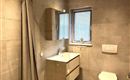 Ein modernes Badezimmer mit hellen Fliesen und einer Dusche. Es gibt ein Waschbecken mit einem Spiegel und ein Fenster mit Jalousien.