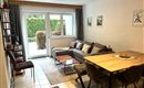 Ein modernes Wohnzimmer mit einer grauen Couch und einem Esstisch. Durch große Fenster scheint Tageslicht und der Blick geht auf einen grünen Garten.