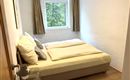 Ein gemütliches Schlafzimmer mit einem großen Bett und hellen Vorhängen. Der Blick aus dem Fenster zeigt grünes Laubwerk.