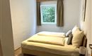 Ein gemütliches Schlafzimmer mit einem Doppelbett und frischen Bettwaren. Das Zimmer hat ein Fenster mit Blick auf Bäume und helle Wände.