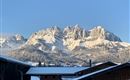 A snow-covered mountain range under a clear blue sky. In the foreground, there are buildings that surround the rural landscape.