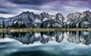An impressive mountain landscape with snow-capped peaks is reflected in the calm water of a lake. The sky is cloudy, giving the scene a dramatic atmosphere.