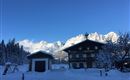 A picturesque winter landscape with snow-covered mountains in the background. In the foreground, there is a traditional alpine cabin and a wooden shed.