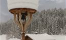 A lantern stands on a snow-covered roof, surrounded by a winter landscape. The trees in the background are also wrapped in snow, while the sky is cloudy.