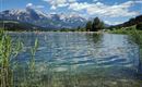 Ein klarer See umgeben von Wiesen und Bergen. Der Himmel ist blau und mit einigen Wolken versehen.